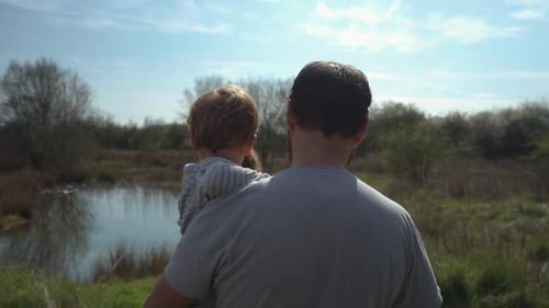 Father walks and carries toddler boy in field by lake, slow motion