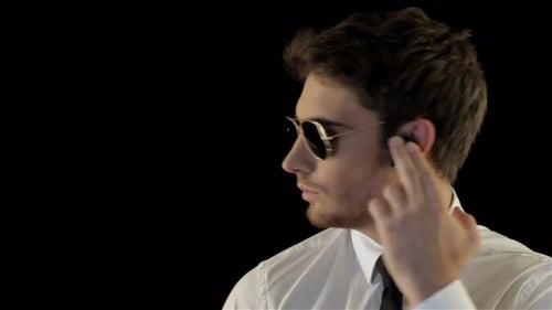 Man Taps Wireless Earpiece Wearing Sunglasses and Tie