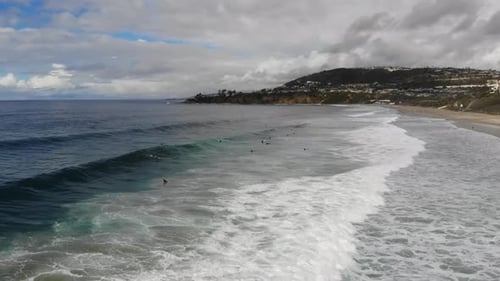 A drone glides low at the beach revealing crashing waves and surfers surfing.