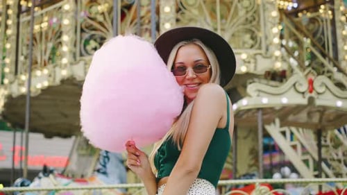 A Young Blonde Woman in a Hat and Sunglasses Holds Cotton Candy Smiling in Front of a Glowing