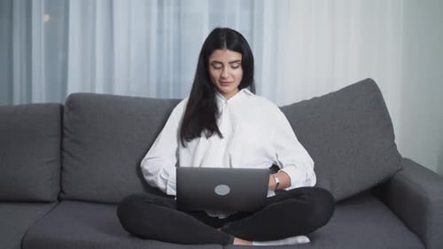 Young Woman Freelancer is Sitting on the Couch and Working on a Laptop Types Text on the Keyboard