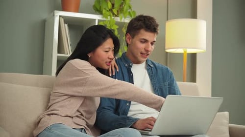 Young Multiracial Couple Sitting on the Sofa at Home Using a Laptop to Search and Book a Hotel Room
