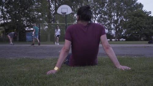 Jeune homme assis sur l'herbe tout en se reposant et regardant des amis jouer au basket de rue