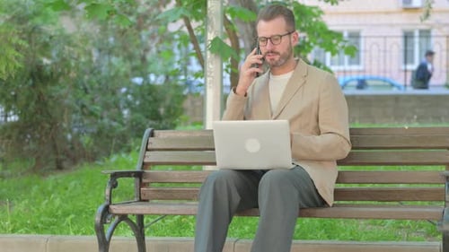 Man Working on Laptop in Park Answers Phone
