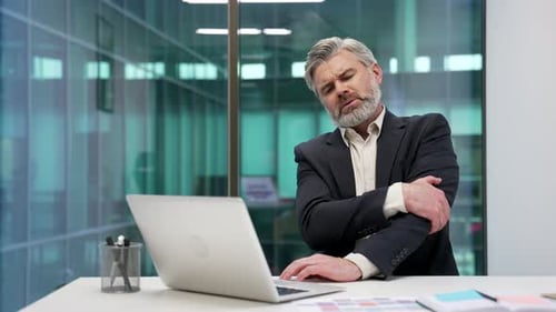 Mature bearded businessman suffering from elbow pain sitting in a business office. Upset male