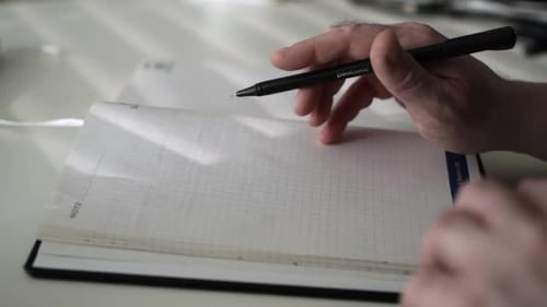 Man Writes in Notebook at Desk With Pen