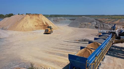 Bulldozer transports sand in a bucket. Asphalt plant surrounded by piles of crushed stones