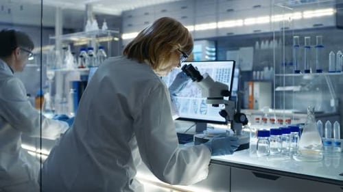 Senior Female Biologist Works on Her Computer and Looks at Materials under Microscope. She's in a M