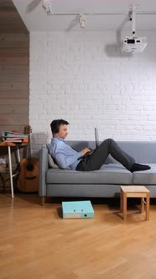 Vertical Video Young Man Wearing Headphones and Working on a Laptop While Relaxing on a Comfortable