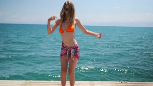 Handsome girl dancing to music, smiling in sunglasses in bright swimsuit ocean background.