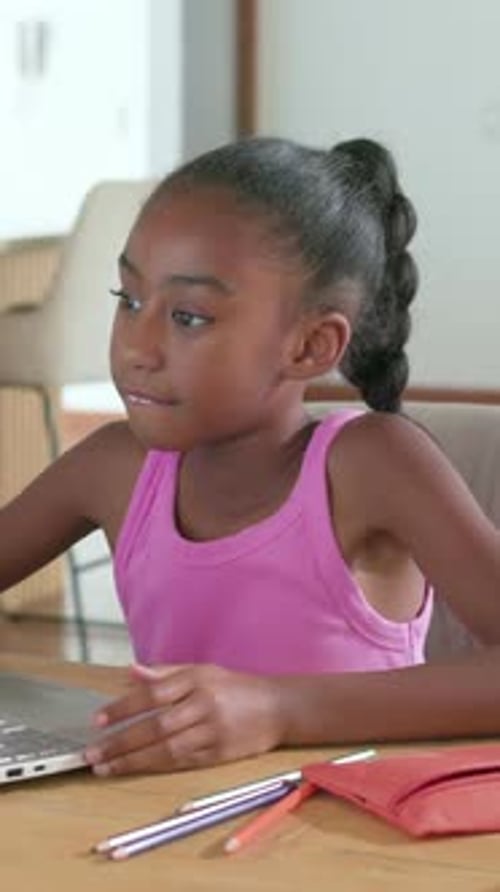 Vertical video: Glancing left, child in pink top using laptop pad at table, learning with pencils
