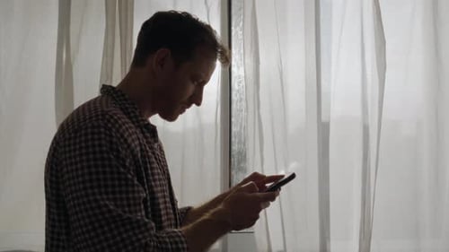 Texting on smart phone. Man standing indoors by window with white curtains, focused on phone. Soft d