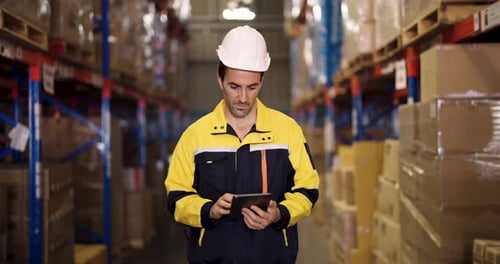 Warehouse worker in industrial safety uniform and helmet holds digital tablet while inspecting cargo