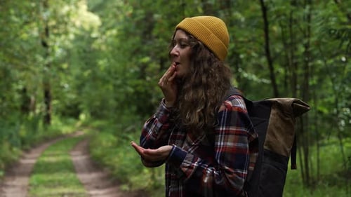 Young Hiker Woman with a Backpack and Yellow Beanie Enjoys Eating Wild Berries Found in a Lush
