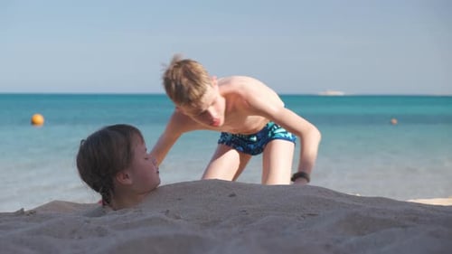 Two Kids Play on Beach Laughing Boy Covers Smiling Girl with White Sand Bright Blue Sky and Sea