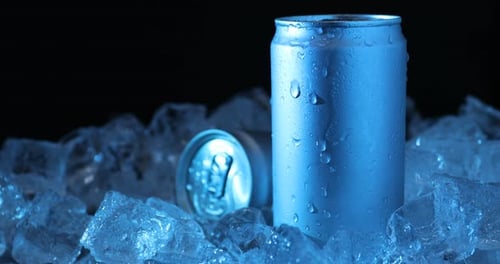 Cans of refreshing drink and ice cubes in blue light on black background, closeup
