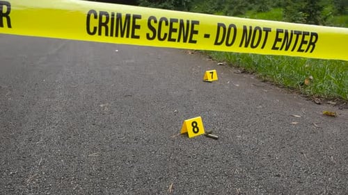 Yellow crime scene tape in front of crime markers and high caliber bullets on asphalt ground outside