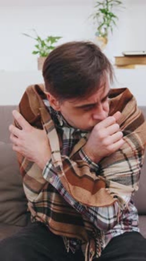 Vertical Video Senior Man Sitting on Couch Wrapped in a Blanket Shivering and Coughing in a Cozy
