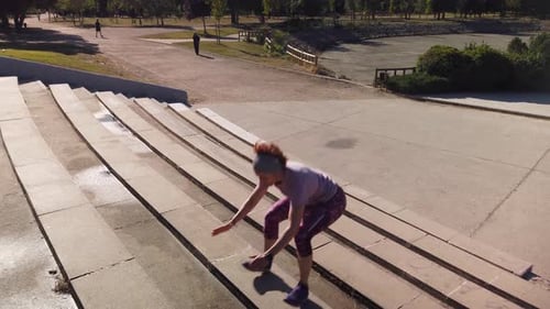 A shot focused on a redhead woman jumping some bleachers in a park.