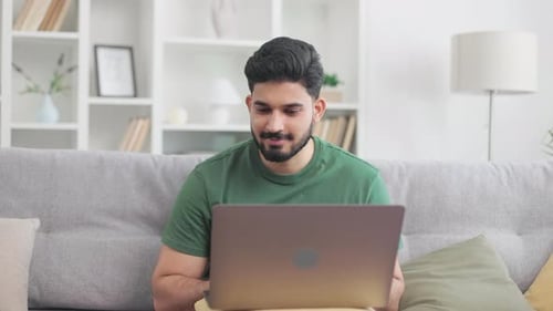 Homme hindou positif assis sur le canapé et tapant sur un ordinateur portable