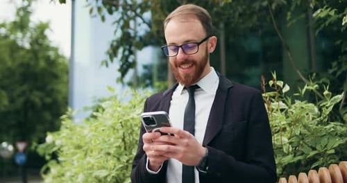 Young smiling businessman is using a smartphone while sitting on a bench near an office building.