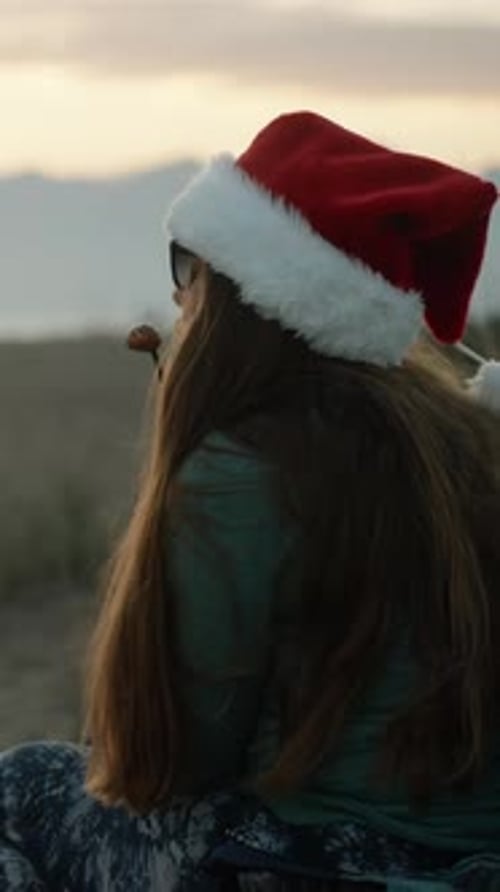 Vertical video. A young woman in a Santa hat against the backdrop of mountains and a sunset offers y