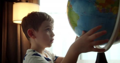 Child Sitting at a Table at Home in Soft Sunlight Carefully Looking at the Globe Rear View of a