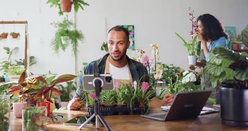 Apuesto jardinero en delantal usando el teléfono y grabando un vídeo de blog con los clientes. Contenido para adultos jóvenes