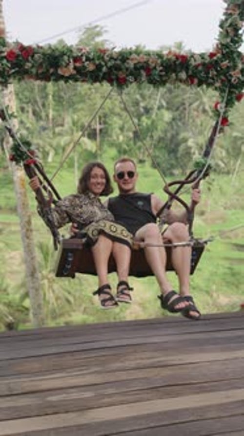 Loving couple on flowers decorated swings in Bali jungle forest, front vertical view