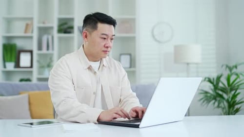 Confident concentrated asian man typing on laptop while sitting at desk at workplace in home office.