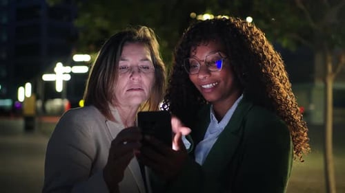 Two Women Looking at Phone in Urban Area at Night