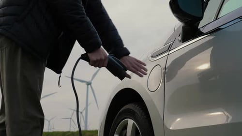 Man Charging Electric Car with Wind Turbines in Background