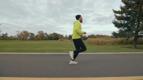 Side View of Sporty Man Running in Autumn Park Listening to Music in Earphones