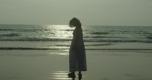 Indian woman stands on the beach in a white dress. The sky is cloudy and the sun is setting in India
