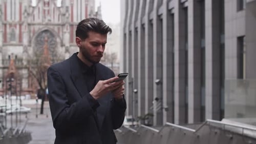A Serious Young Man in a Black Suit is Standing on the Street and Typing Something on a Smartphone