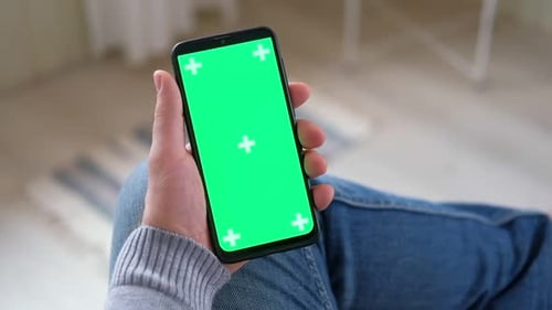 Young Man Sitting at Home Holding Smartphone Green Mockup Screen in Hand Male Person Using Chroma
