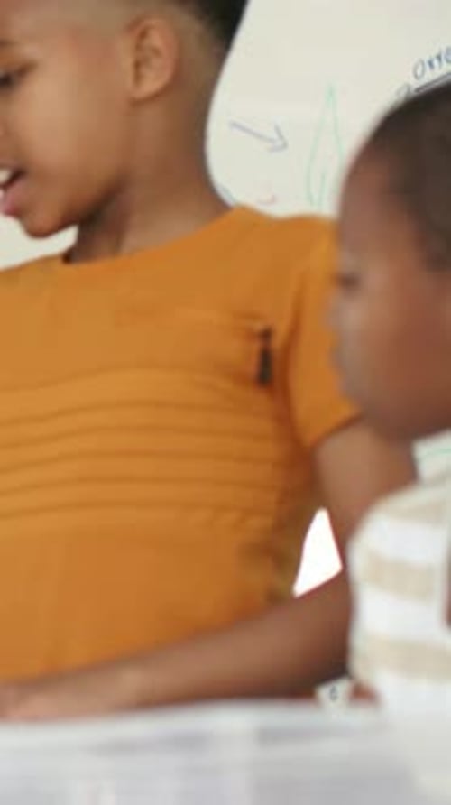 Vertical video: Turning head, kid in orange rising, sharing OXYGEN in class at table with board bin