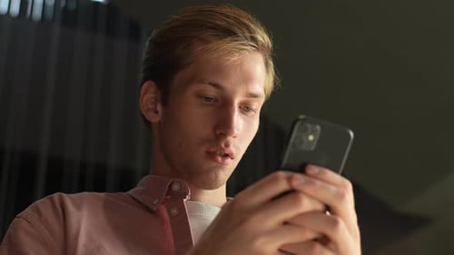 Closeup Lowangle View of Serious Young Man Browsing Internet on Mobile Phone Sitting on Sofa at Home