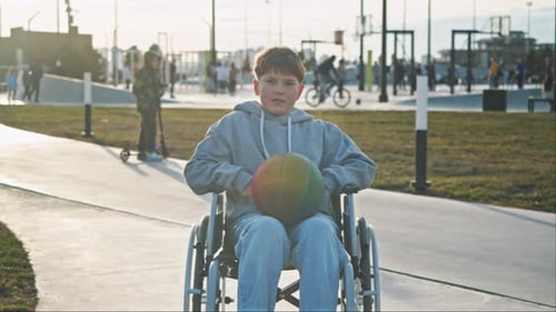 A Teenage Boy with Special Needs is Sitting in a Wheelchair in a Public Park Looking Around