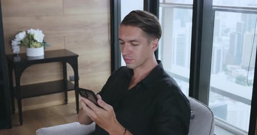 Businessman in Black Shirt Uses Mobile Phone Sitting in Chair at Office on Window Background