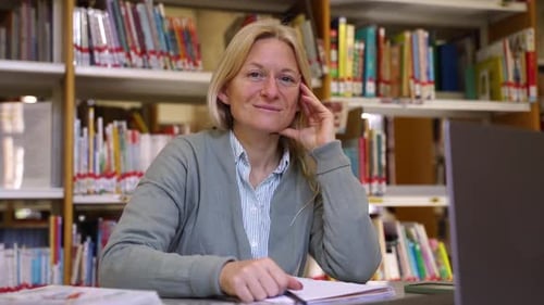 Mature Teacher Woman Looking at Camera Over School or College Library Book Shelf and Education