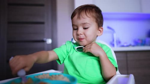 Baby boy eating slowly with a spoon. Adorable toddler in light green t-shirt having meals.