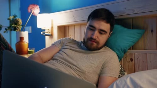 A Young Man Wearing Casual Clothing is Using His Laptop in a Cozy Bedroom at Night for Relaxation