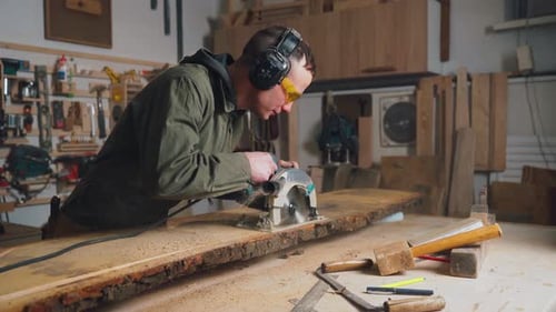 Worker Male Builder Works in a Workshop Cuts a Board with a Circular Saw in Goggles and Headphones