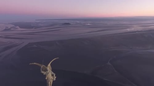 Statue of Saint Michael on spire of Mont Saint Michel abbey tower with bay in background at sunset.