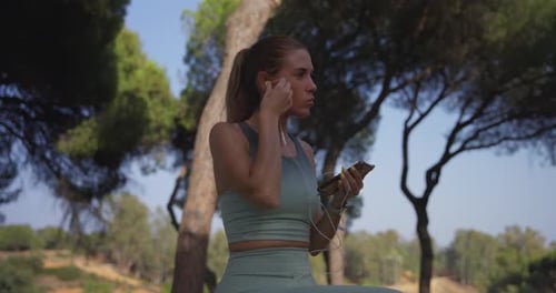 Blonde Woman Puts in Earbuds in a Green Park