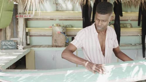African american man preparing surfboard on the counter of surf rental beach shack, slow motion