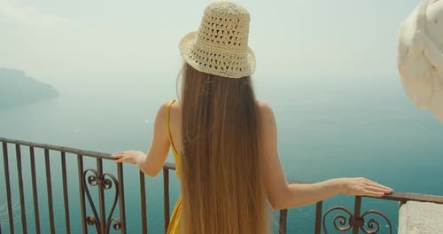 A woman enjoys a serene view of the sea from a balcony in Italy, framed by ornate ironwork. Tourist