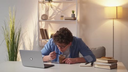 Young Man Eats Noodles at Laptop Desk