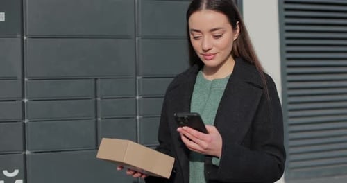 Woman Receiving Parcel From Post Terminal Machine Using Smartphone Outdoors